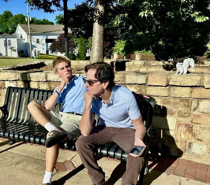 Two people sit on a bench near a low stone wall with a white squirrel sculpture on top.