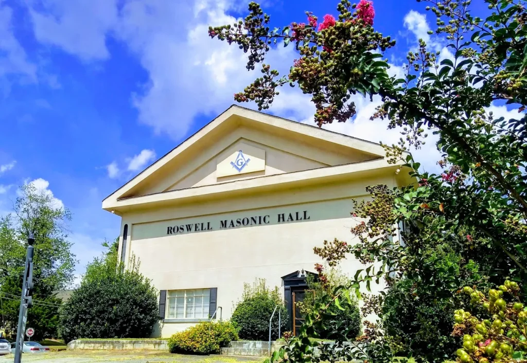 The front of Roswell Masonic Hall framed by trees and flowers under a blue sky.