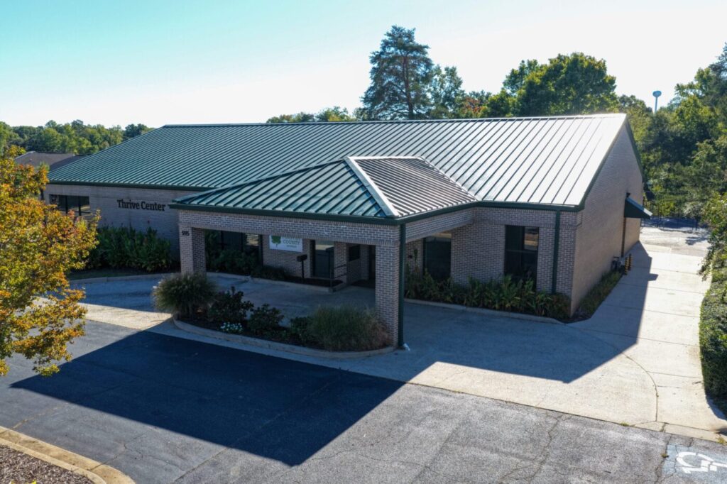 A single-story brick building with a green metal roof and a covered entrance surrounded by trees
