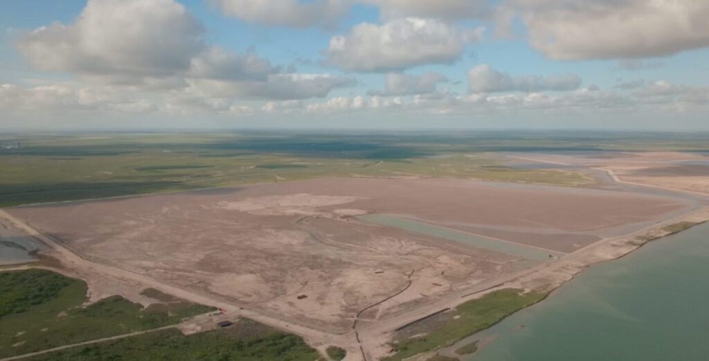 Aerial view of a broad cleared construction site beside water under a cloudy sky.