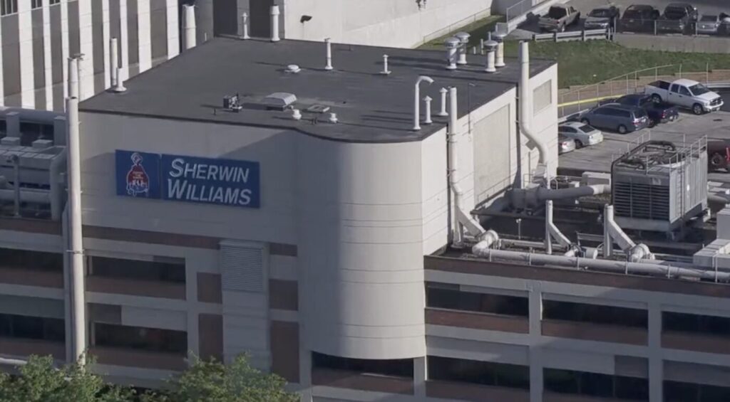 Sherwin-Williams sign on a large industrial building with rooftop equipment and parked cars nearby.