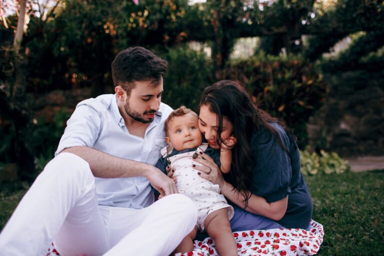 Two adults sit on grass with a baby between them outdoors.