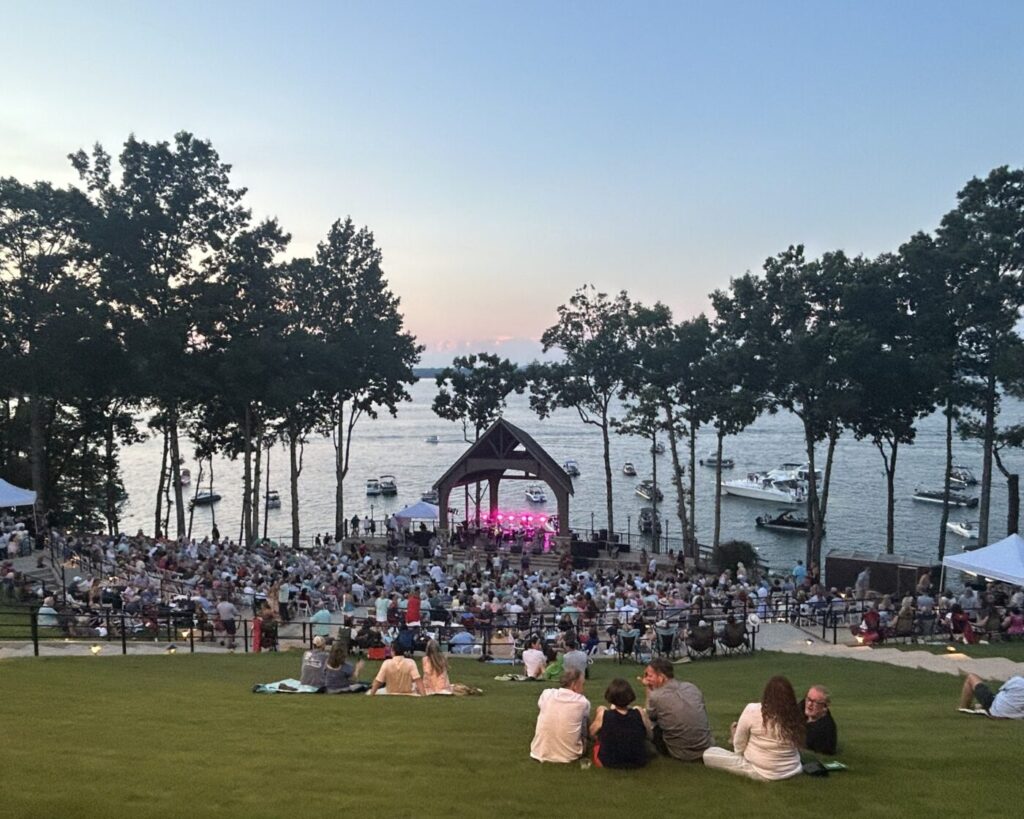 An audience watches a concert at a lakeside amphitheater, with boats on Lake Lanier behind the stage.