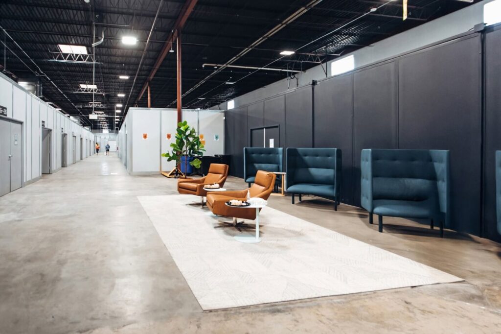 Warehouse corridor with private suite doors, lounge chairs and small tables on a light rug.