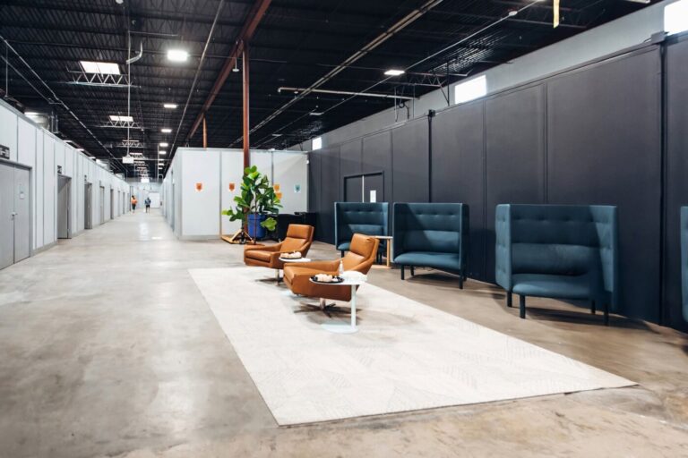 Warehouse corridor with private suite doors, lounge chairs and small tables on a light rug.