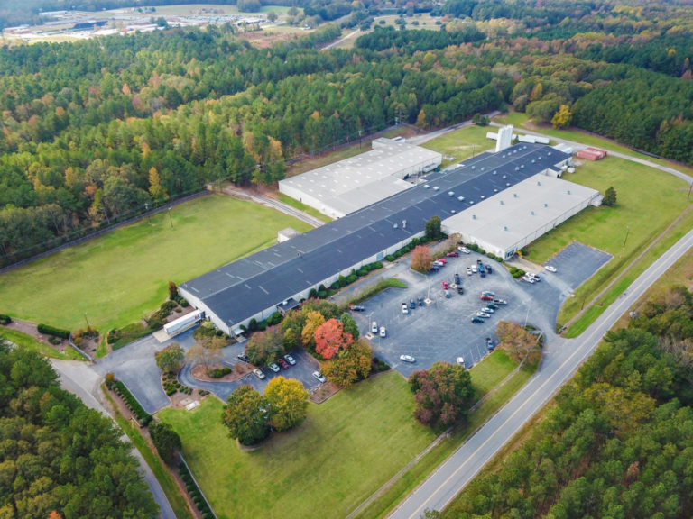 Aerial view of a large industrial building surrounded by trees, with parking lots and access roads.