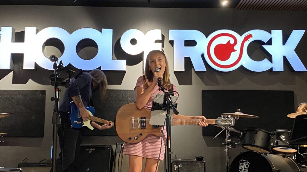 A young singer holds a guitar and sings into a microphone while another musician plays guitar beside her.