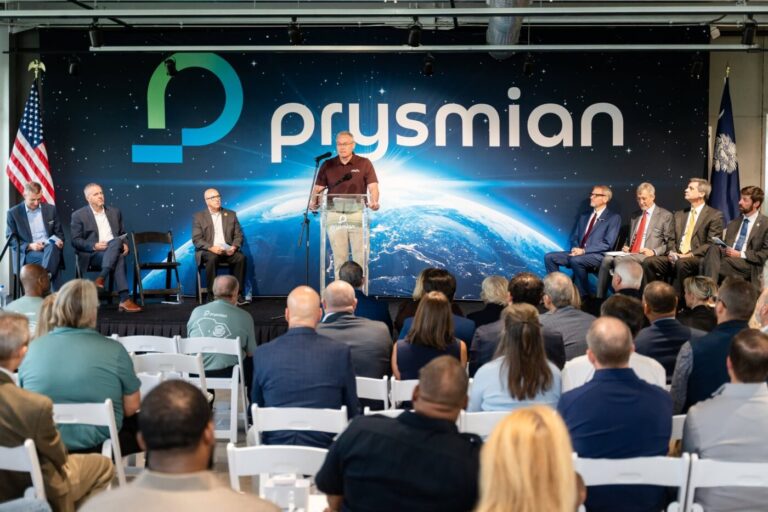 A man speaks at a podium in front of a Prysmian backdrop as seated attendees listen.