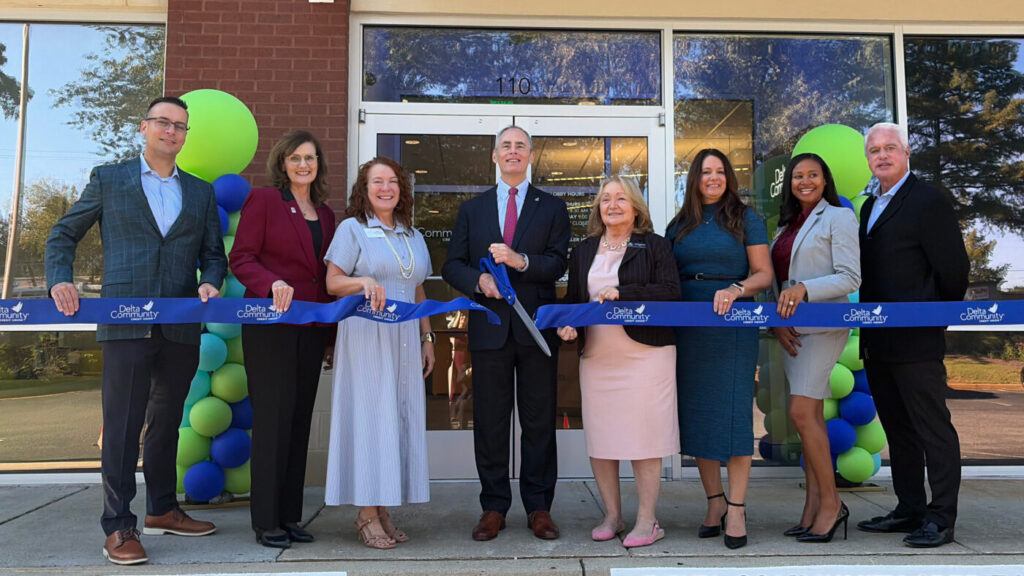 Eight adults stand outside a credit union branch holding a blue ribbon during a ribbon-cutting ceremony, with green and blue balloons beside the entrance.