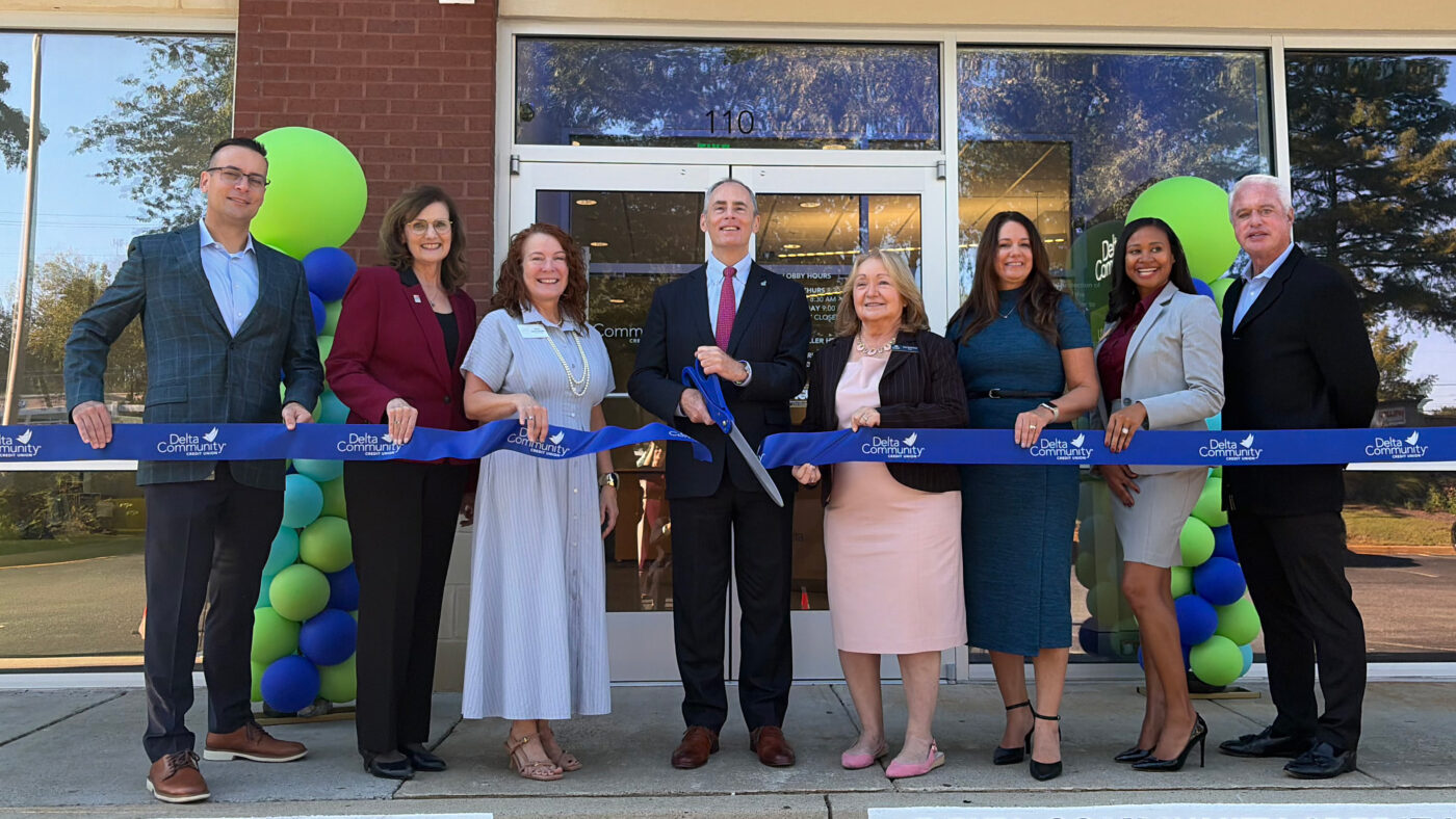 Eight adults stand outside a credit union branch holding a blue ribbon during a ribbon-cutting ceremony, with green and blue balloons beside the entrance.