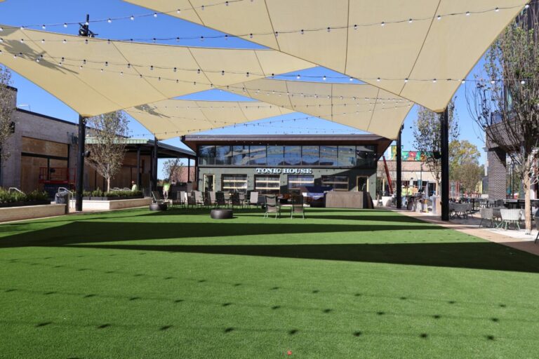 Shade sails above a green plaza with chairs, workers and Tonic House in the background.