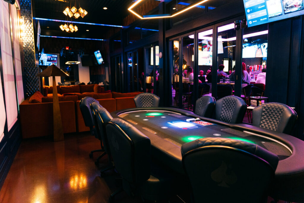 A card table sits empty in a dark, neon-lit room with lounge seating and screens in the background.