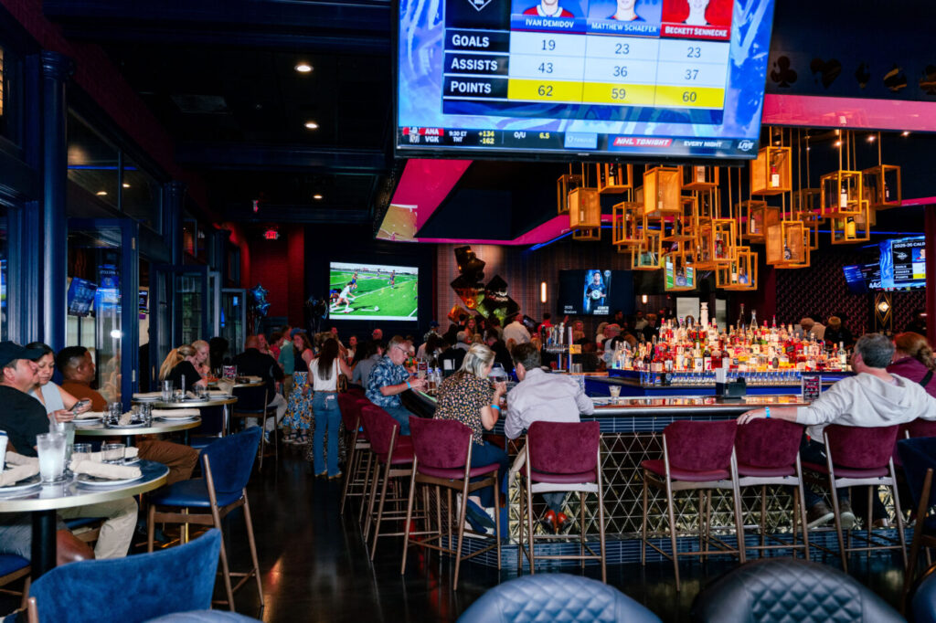 A busy bar and dining area with patrons seated at tables under large TV screens and hanging lights.