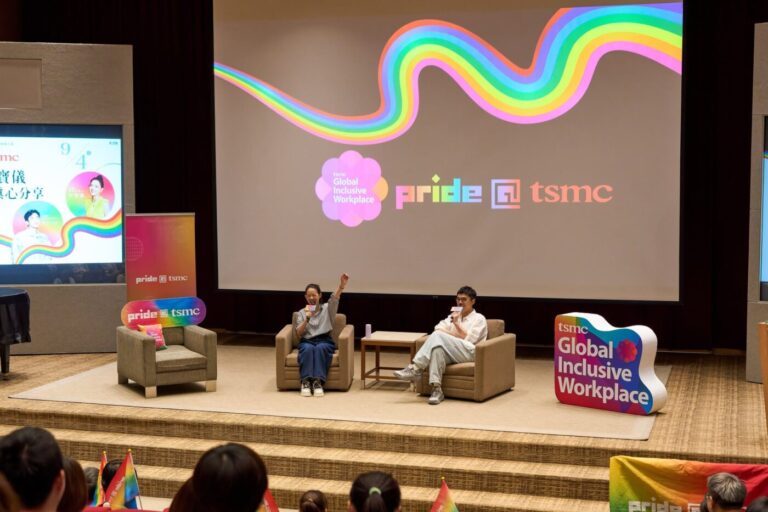Two speakers seated on chairs on a stage in front of a large screen with a rainbow Pride@tsmc graphic