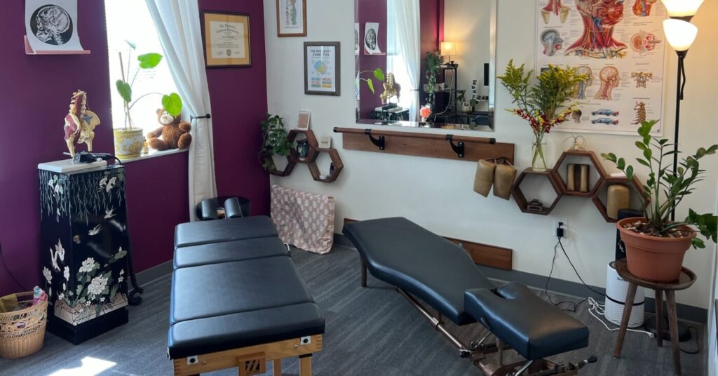 A treatment room with a massage table, adjustable bench, wall anatomy posters, framed certificates and potted plants.