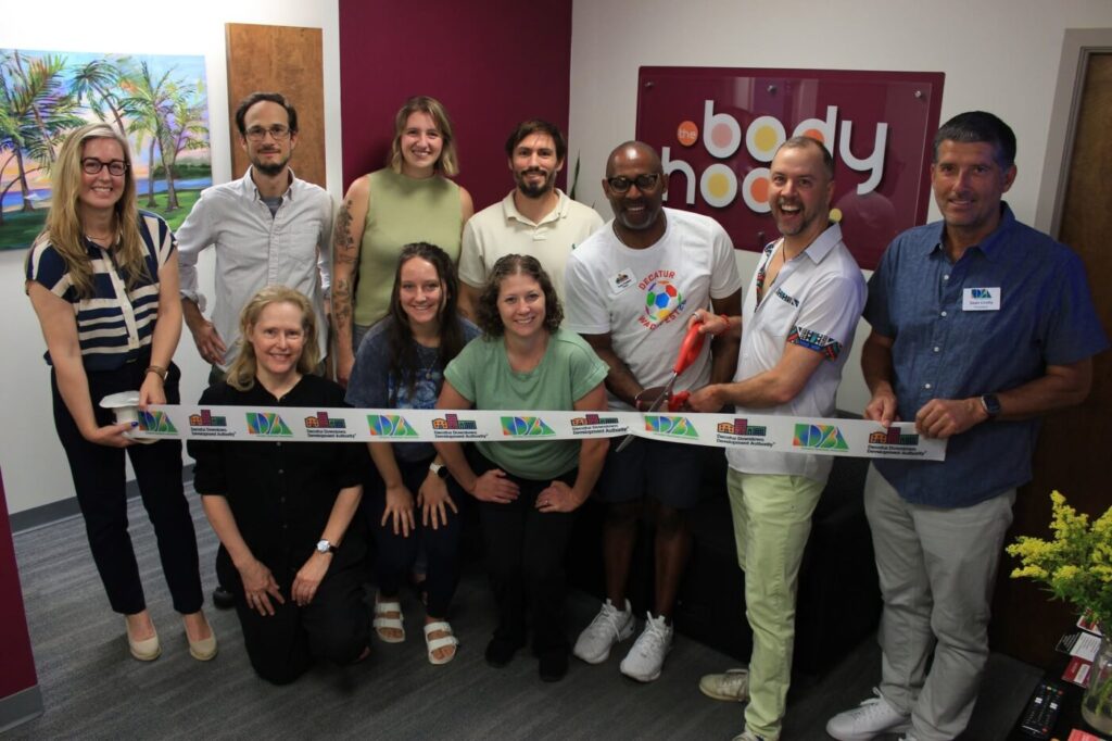 A group of people stands indoors holding a ribbon across a doorway in front of a wall sign for The Bodyhood.