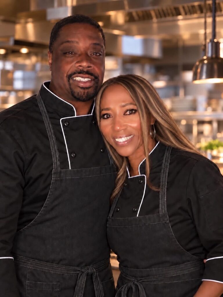 A man and woman in black chef jackets and aprons stand together in a kitchen.