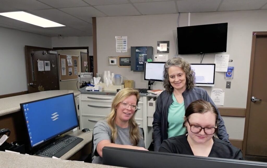 Three women in an office stand and sit behind computer monitors at a nursing facility desk