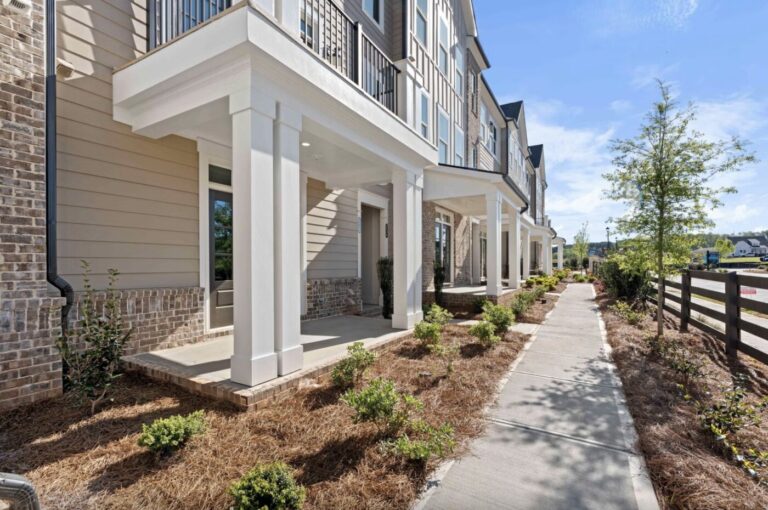 Row of beige townhomes with white porches along a sidewalk under a blue sky