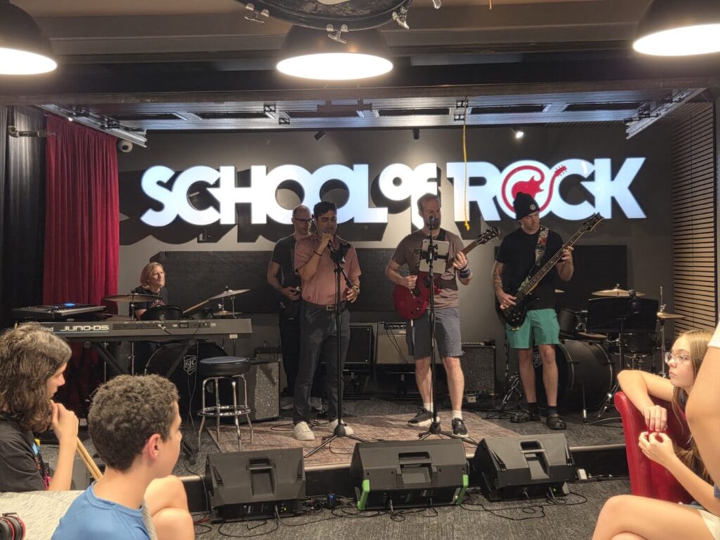 An adult band performs on a small stage while students sit nearby.