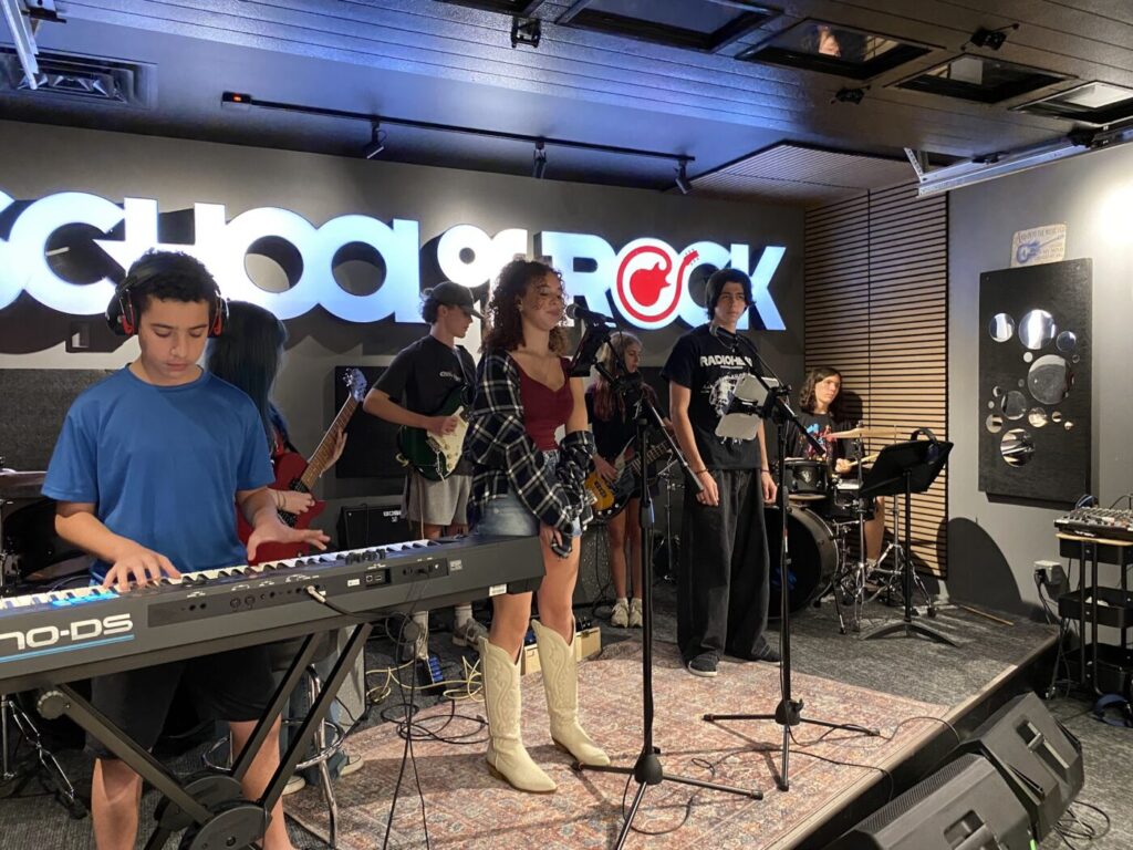 Teen musicians perform onstage with guitars, bass, keyboards and drums.