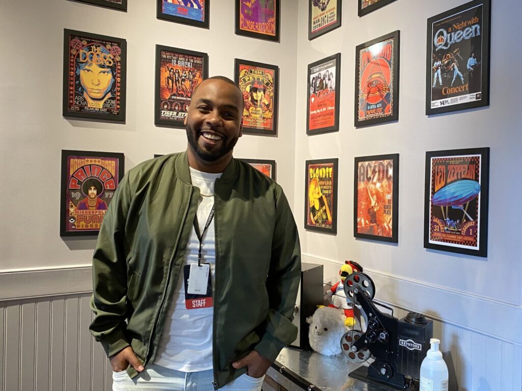 A smiling man stands in a music-themed room lined with framed concert posters.
