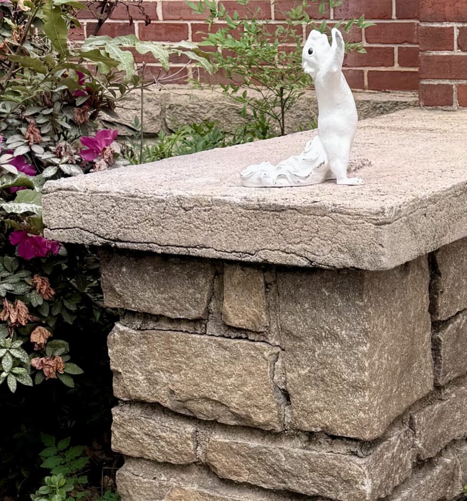 A white squirrel sculpture stands on a stone ledge beside flowers and a brick wall.