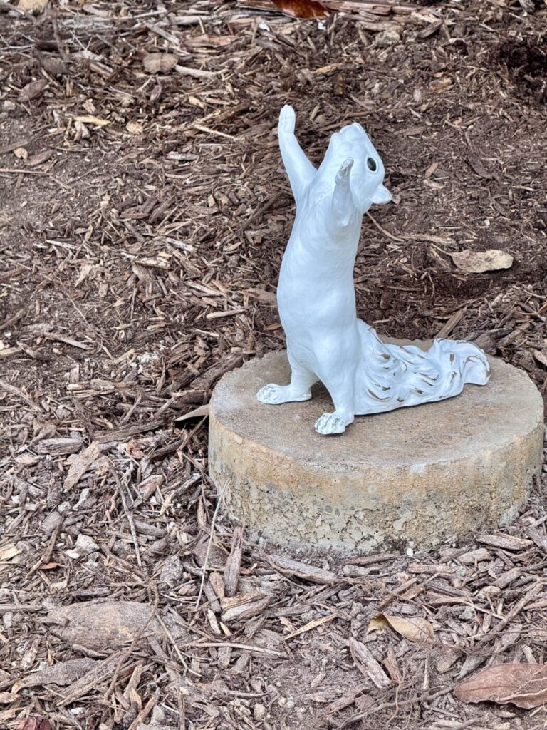 A white squirrel sculpture stands upright with its paws raised on a round base in a mulched area.