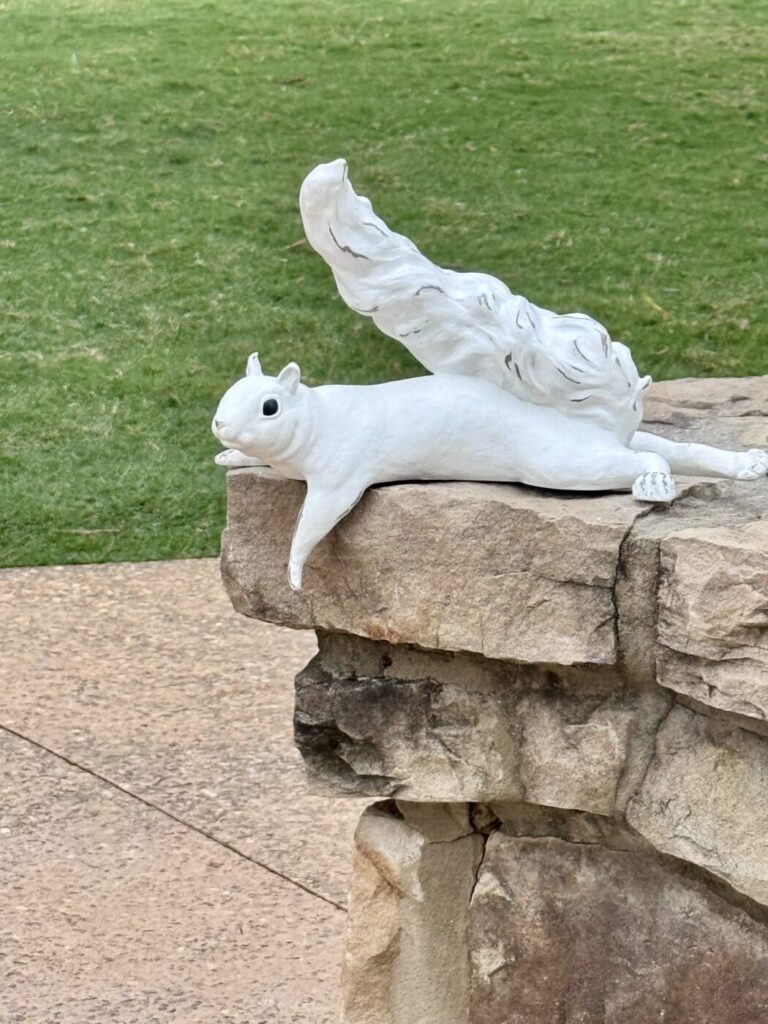 A white squirrel sculpture lies on a stone ledge above a walkway and lawn.
