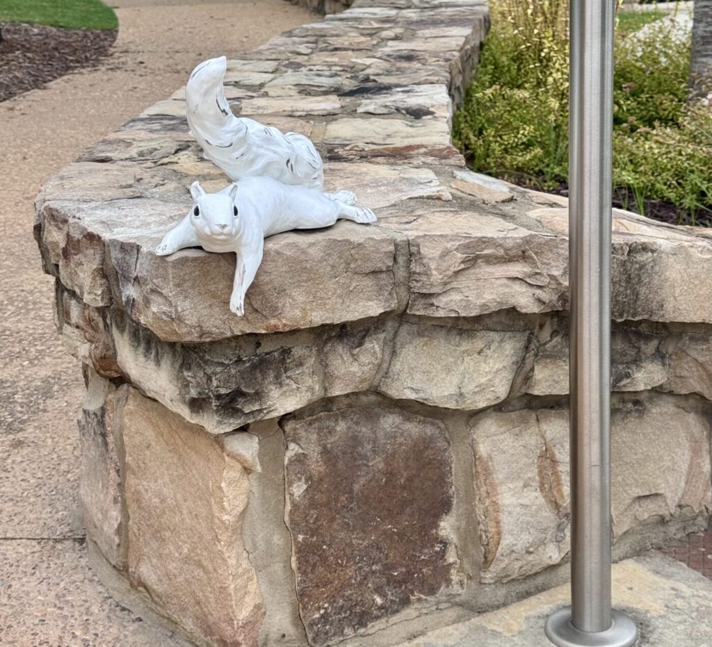 A white squirrel sculpture lies on a stone wall beside a metal post.