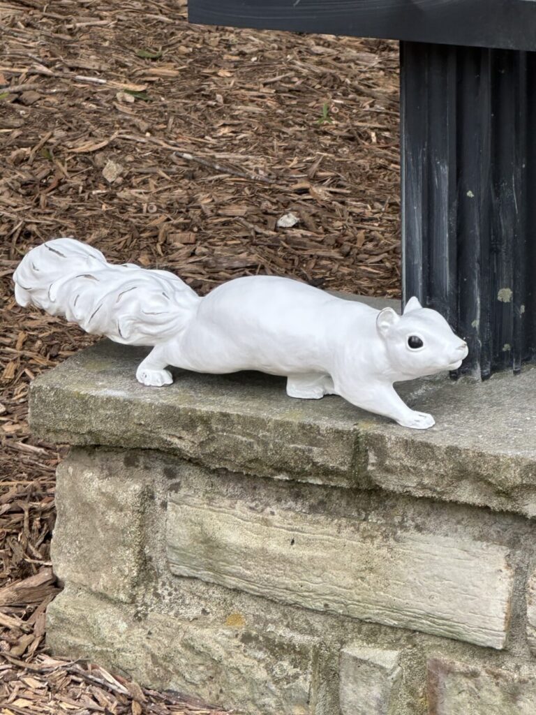 A white squirrel sculpture stretched along the base of a black post in a mulched bed.