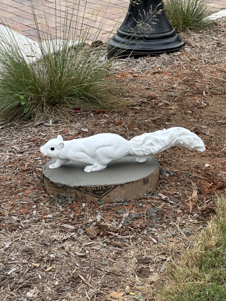 A white squirrel sculpture sits on a circular base near a curb and landscaped verge beside a busy street.
