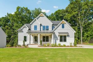 A white two-story house with black roof accents, a covered front porch, and a green lawn in front of trees.
