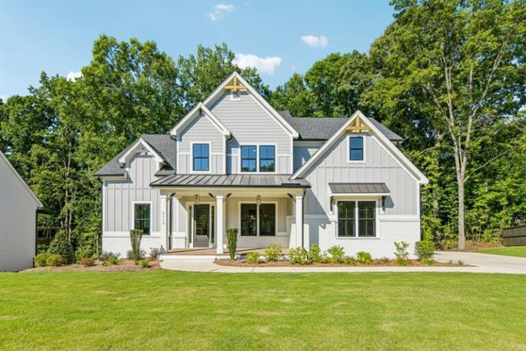 A white two-story house with black roof accents, a covered front porch, and a green lawn in front of trees.