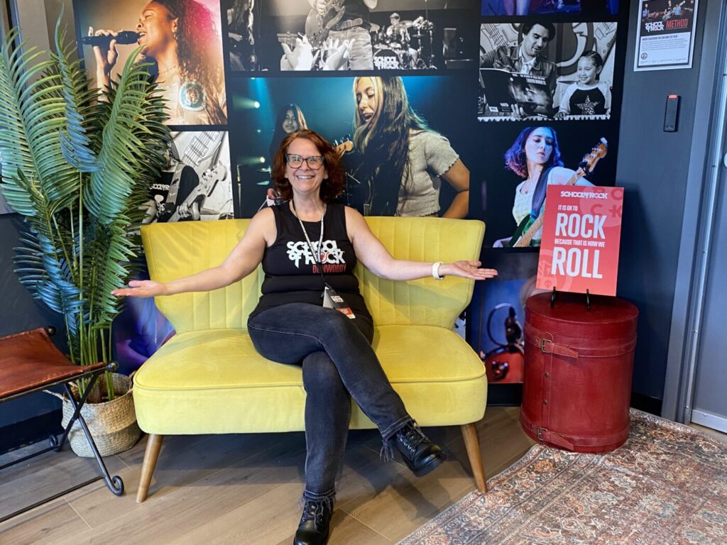 Yael Lindenboim sits smiling on a yellow couch with her arms open in front of a wall of music photos.