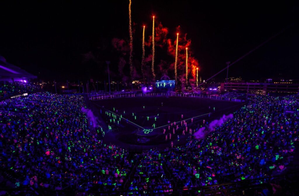 A baseball stadium packed with fans glowing under black lights as fireworks rise above the field at night.
