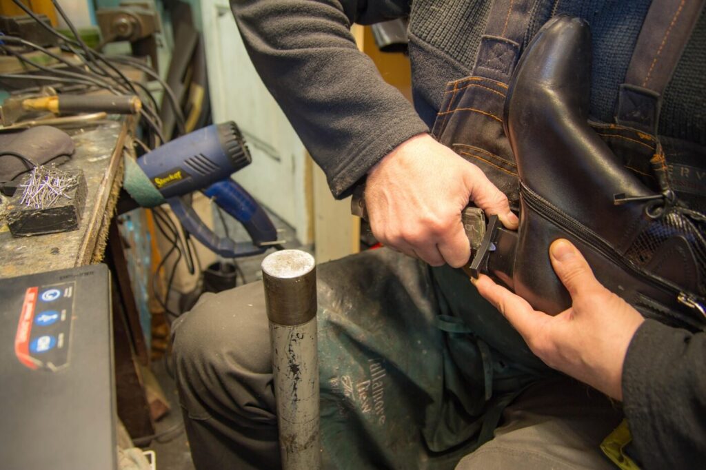 A cobbler trims or repairs the heel of a black boot at a workbench.