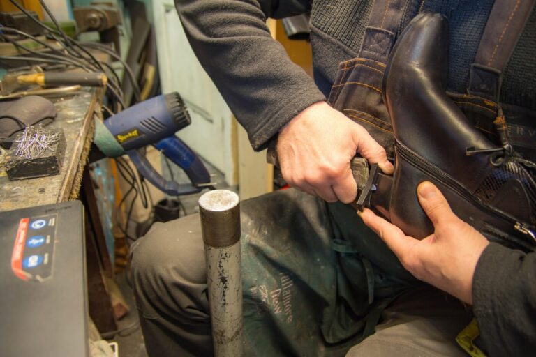 A cobbler trims or repairs the heel of a black boot at a workbench.