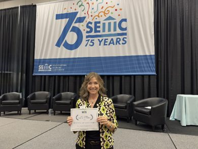 Woman standing on a conference stage holding a certificate in front of a SEMC 75 Years banner and empty chairs.