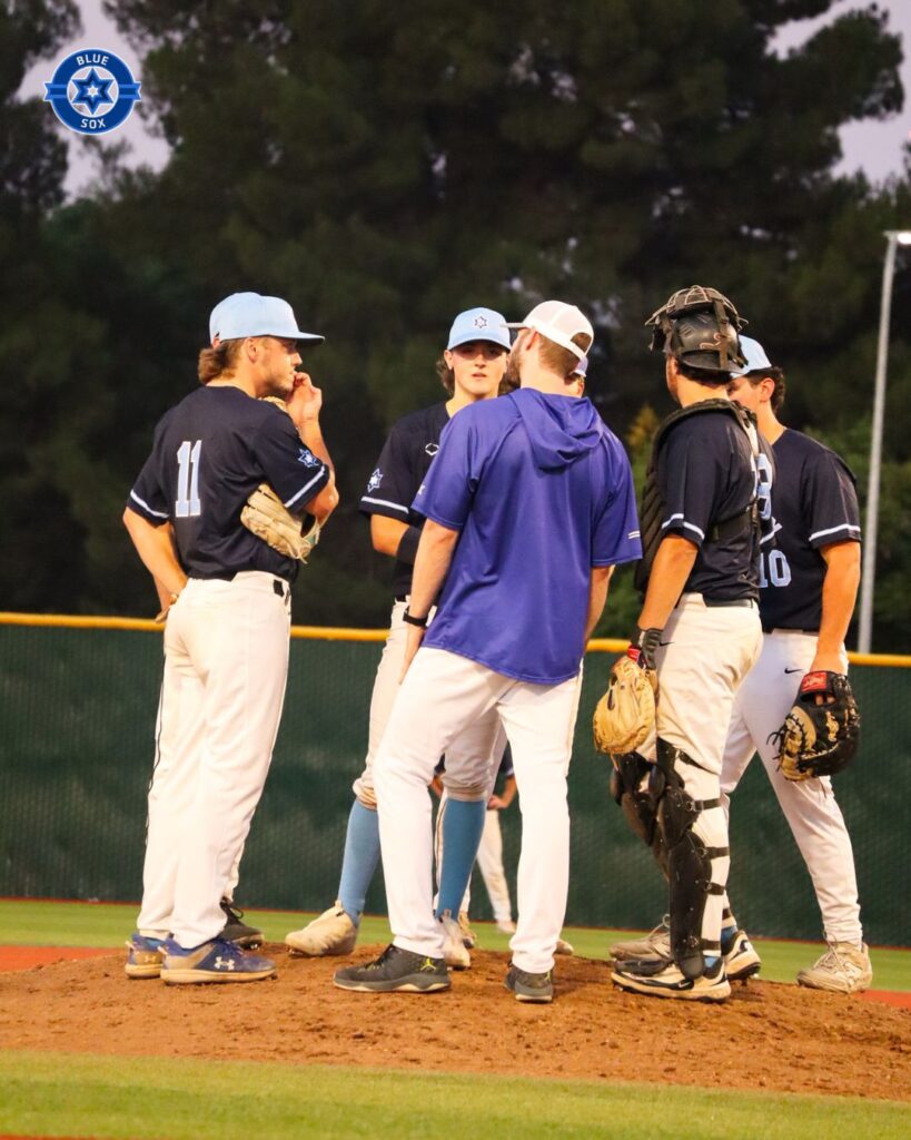 A small group of baseball players and coaches gathers on a mound during an outdoor team discussion.