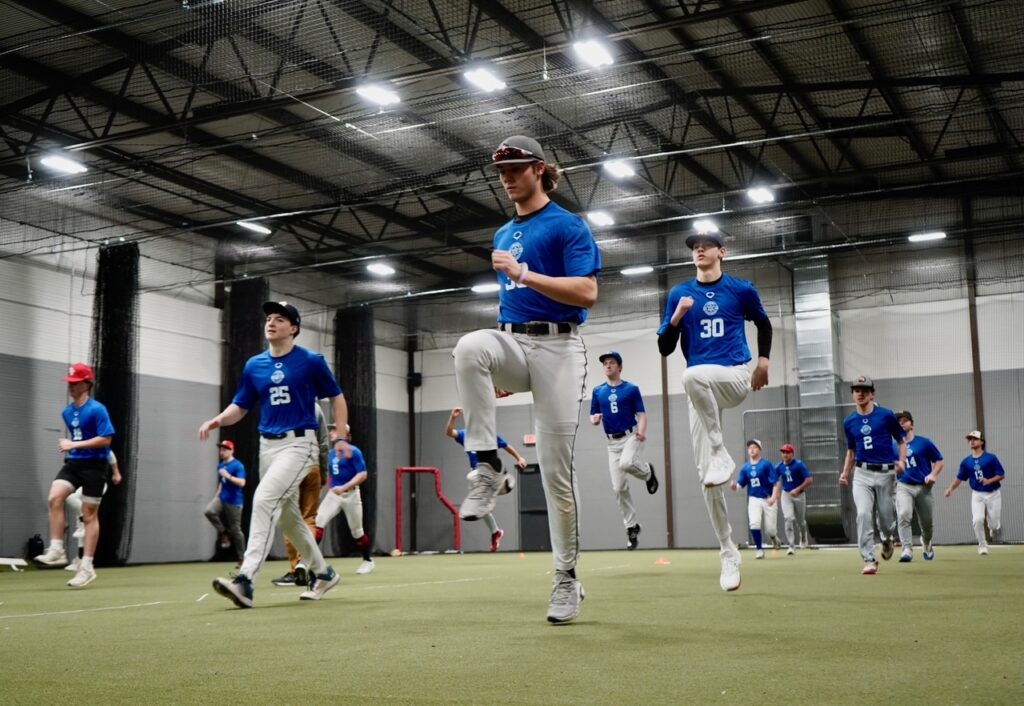 Baseball players in blue shirts and gray pants do conditioning drills inside an indoor training facility.