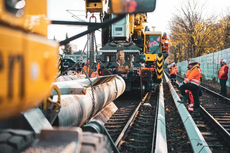 Construction workers in orange safety gear stand beside large rail equipment and suspended pipes on train tracks.