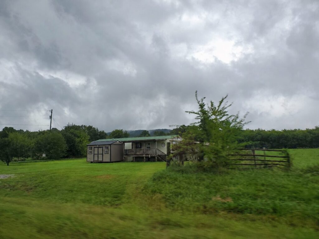 A small house sits beside a shed in a green field under dark storm clouds.