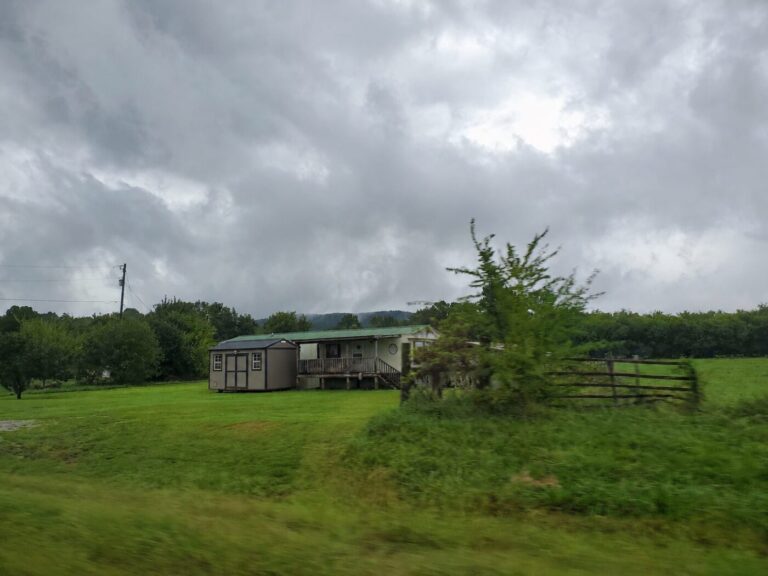 A small house sits beside a shed in a green field under dark storm clouds.