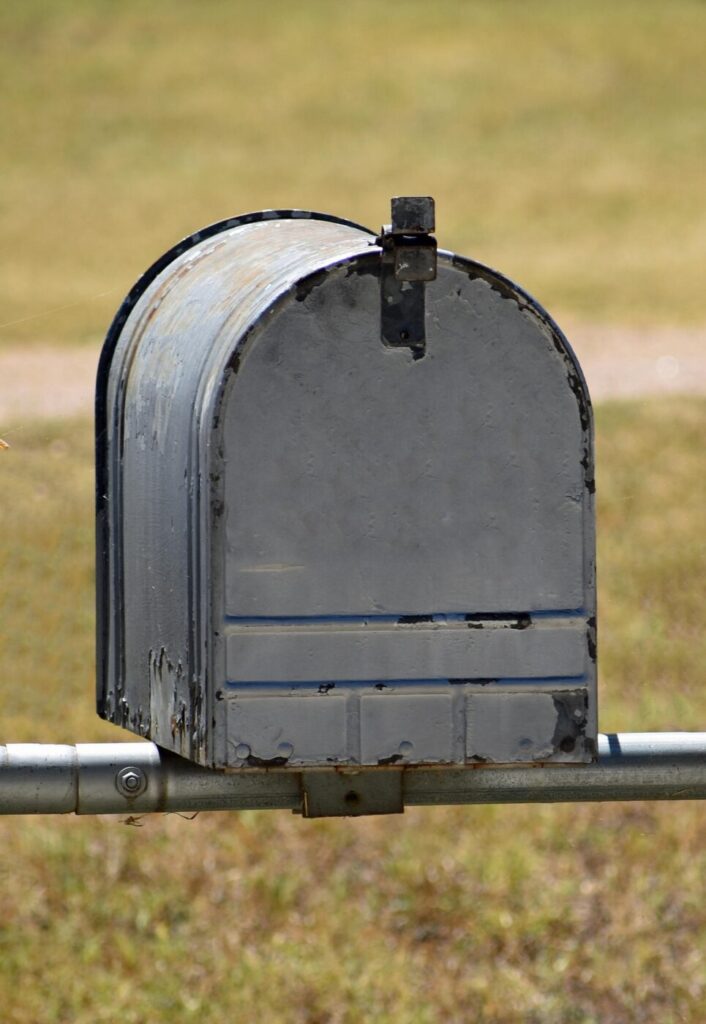 A weathered gray mailbox mounted on a metal post in front of a blurred grassy background.