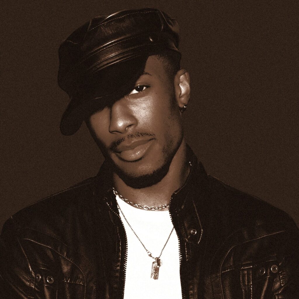 A man in a black leather jacket and cap poses against a dark background.