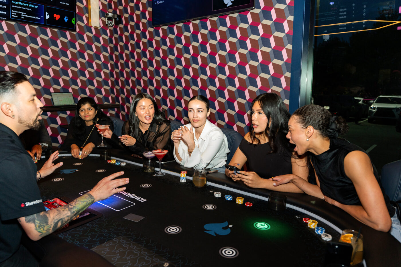 Six adults seated around a curved card table in a dimly lit lounge, with chips, drinks and colorful wall patterns behind them.