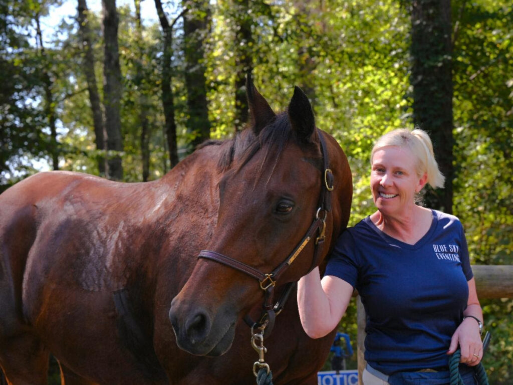 A woman standing beside a brown horse in a wooded area.