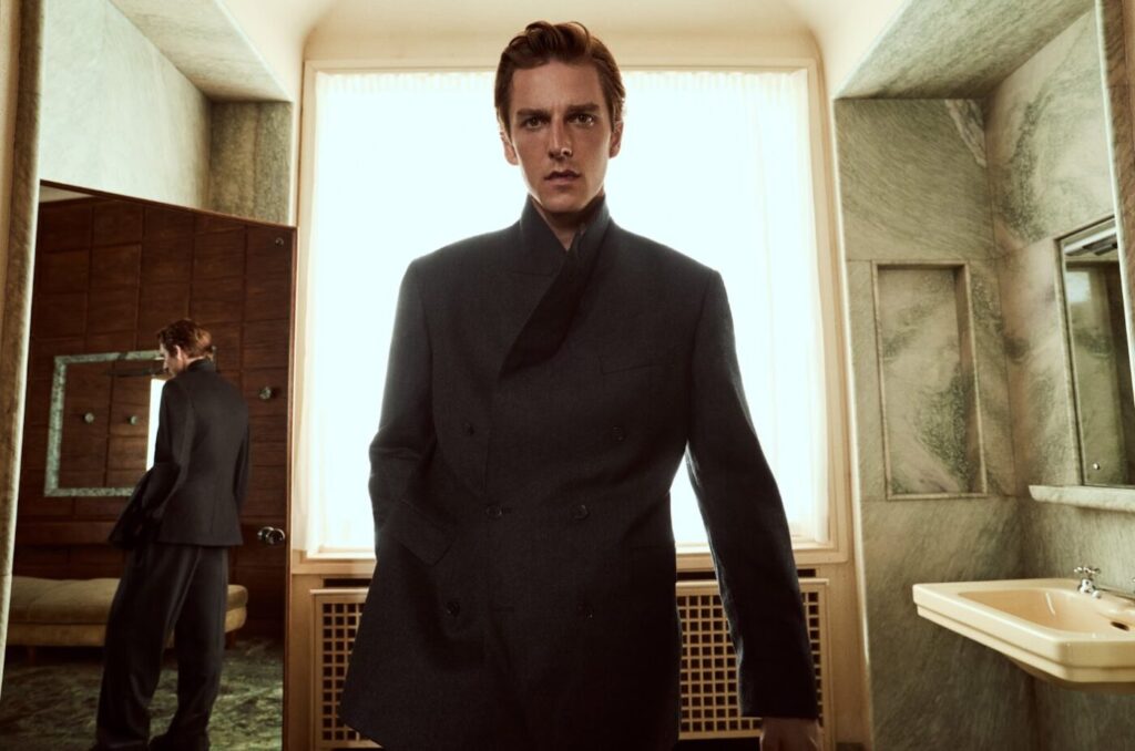 A man in a dark suit stands in a bright room with mirrors and a sink.