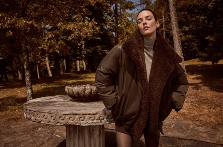 A woman stands outdoors beside a stone table, wearing a dark coat with a wide shearling collar.